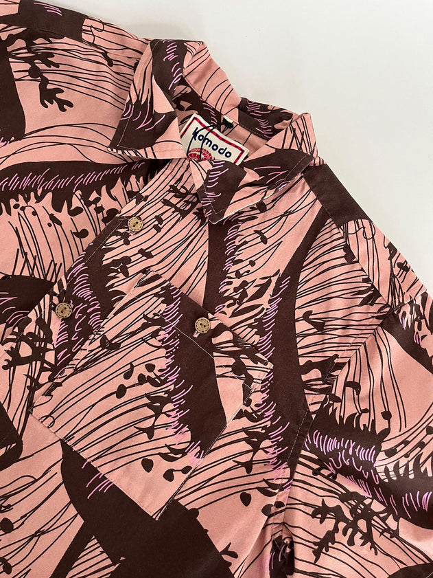 The SPINDRIFT - Organic Cotton Shirt - Pink Wave features a pink and brown abstract wave print, button-up design, Komodo label on the collar, and is laid flat on a white surface.