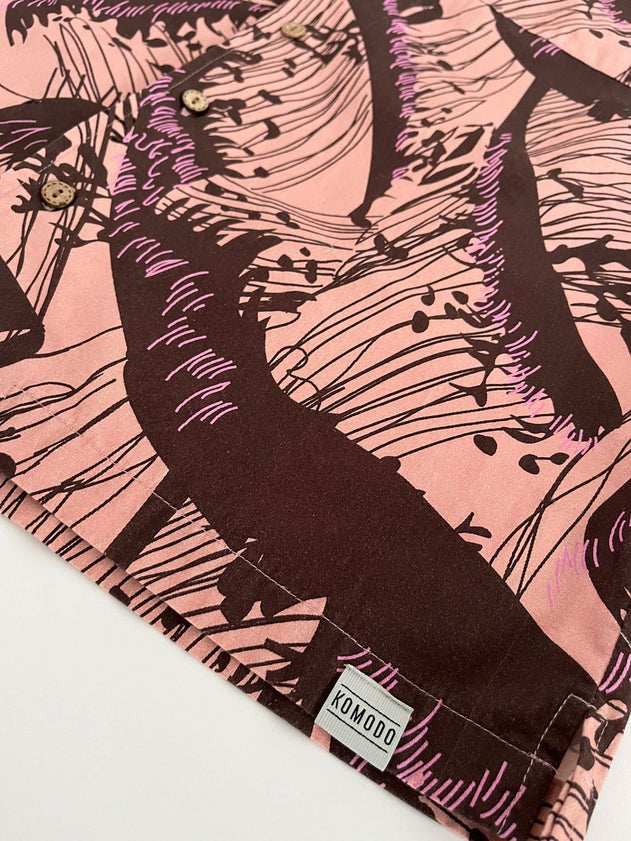 A close-up of the SPINDRIFT - Organic Cotton Shirt - Pink Wave, showing its abstract dark brown and purple wave print, button details, and a small KOMODO fabric tag on the hem.