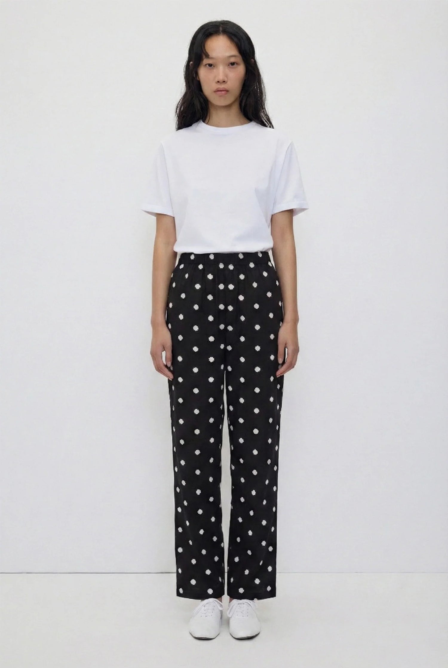 A model stands against a plain white studio background wearing a white short-sleeve crewneck t-shirt and black wide-leg trousers with a white polka-dot pattern, paired with white sneakers.