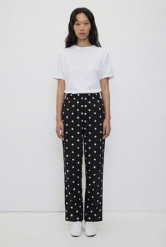 A model stands against a plain white studio background wearing a white short-sleeve crewneck t-shirt and black wide-leg trousers with a white polka-dot pattern, paired with white sneakers.