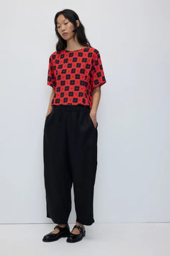 A woman modeling a red short-sleeve top with a black floral pattern, paired with black wide-leg trousers and black Mary Jane shoes, against a plain light gray studio background.