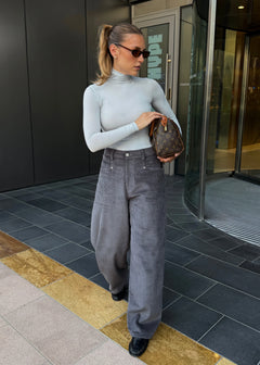 A woman wearing a light gray mock neck long-sleeve top, gray corduroy wide-leg trousers, and black loafers, holding a brown monogram handbag while standing outside a building.
