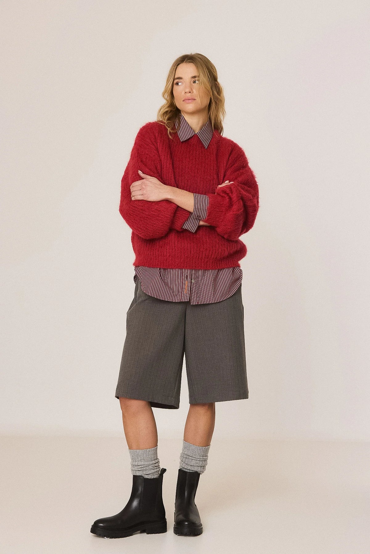 A model stands against a plain light beige studio background wearing a chunky red knit sweater over a gray collared shirt, paired with gray shorts, gray socks, and black boots.