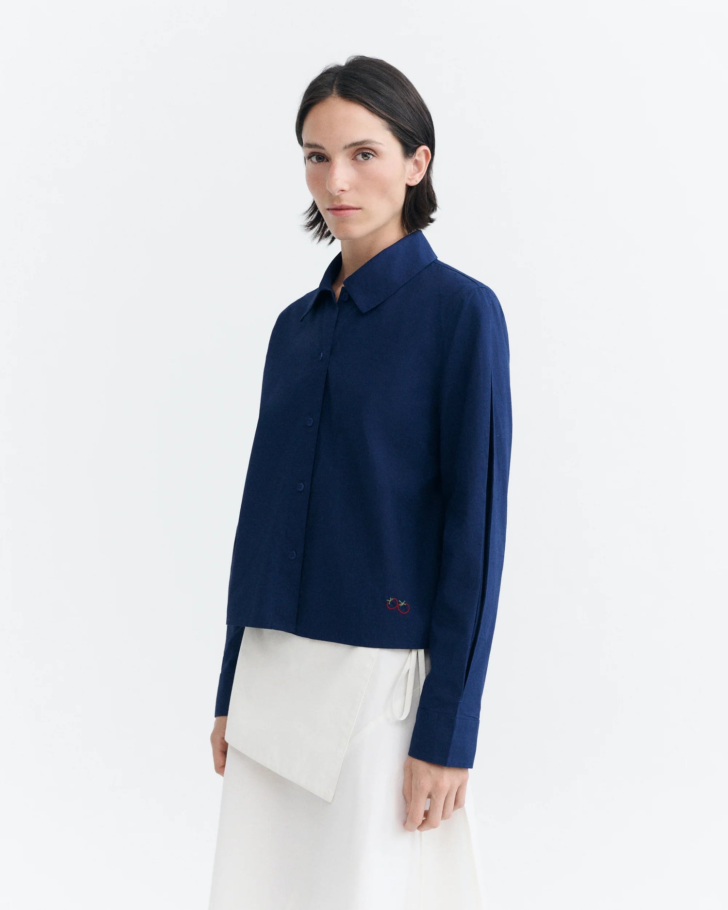 A model wearing a navy blue long-sleeve button-up shirt with a small embroidered logo on the lower left hem, paired with a white asymmetrical skirt, against a plain white studio background.