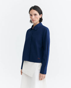 A model wearing a navy blue long-sleeve button-up shirt with a small embroidered logo on the lower left hem, paired with a white asymmetrical skirt, against a plain white studio background.