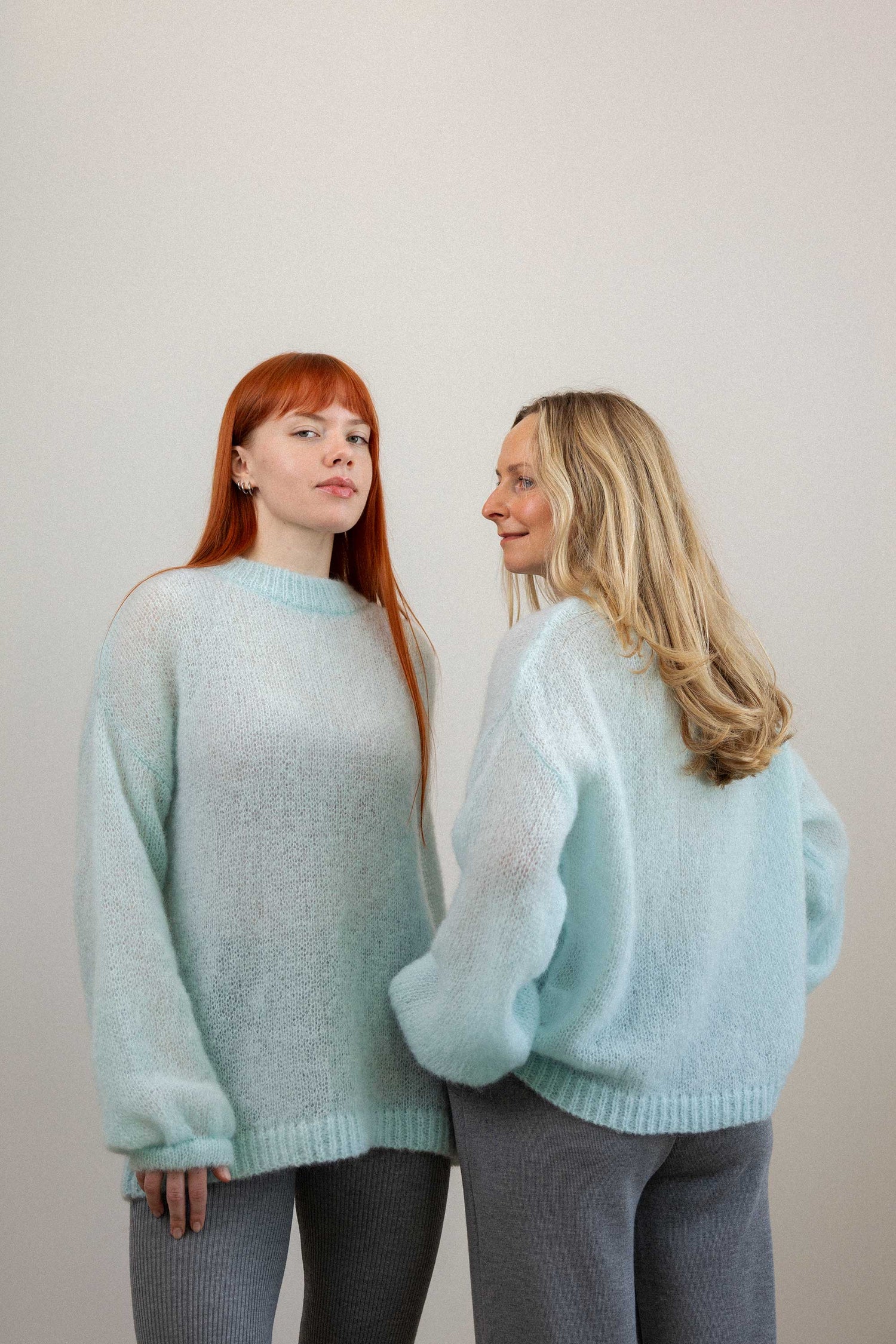 Two women wearing light blue sweaters standing back-to-back against a plain background
