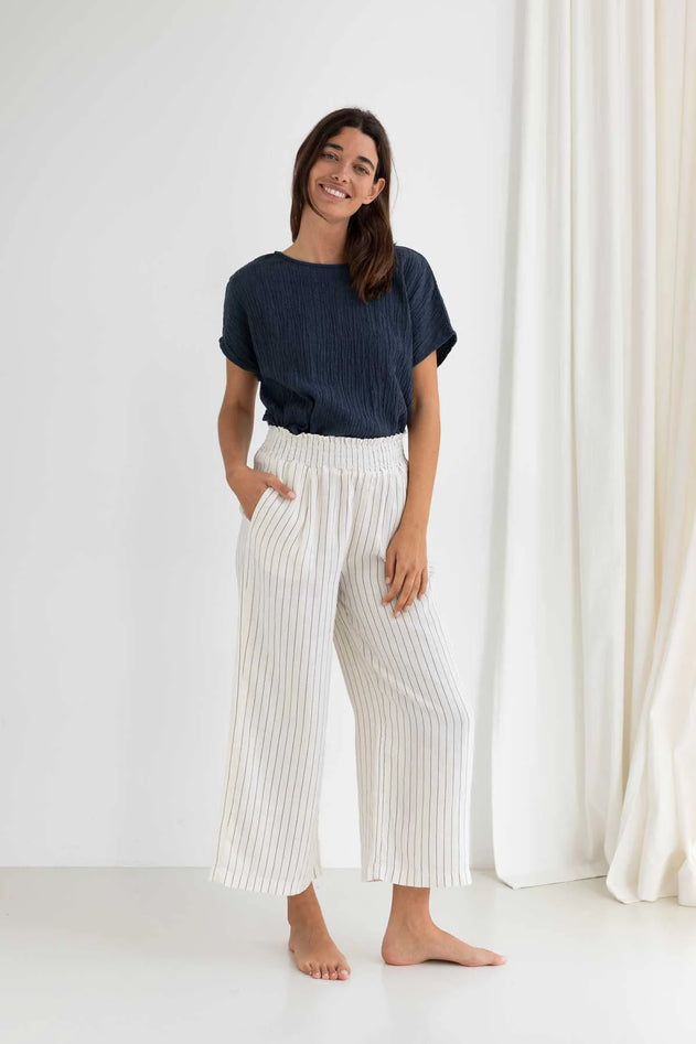 Woman wearing a navy top and white striped pants standing against a white curtain.