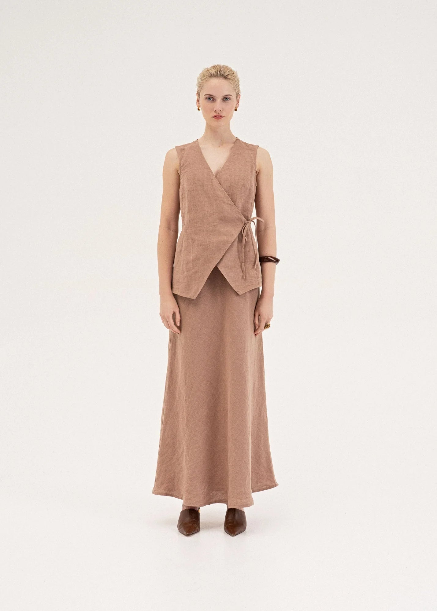 A model stands against a plain light gray studio background wearing a matching two-piece outfit consisting of a sleeveless wrap-style vest and a long skirt in a muted dusty rose or taupe-brown tone, paired with dark brown pointed shoes.