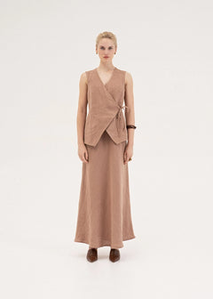 A model stands against a plain light gray studio background wearing a matching two-piece outfit consisting of a sleeveless wrap-style vest and a long skirt in a muted dusty rose or taupe-brown tone, paired with dark brown pointed shoes.