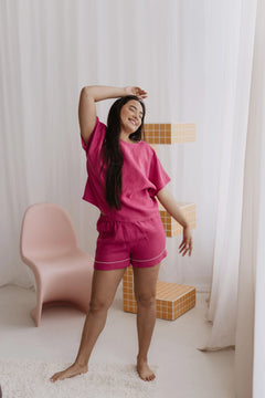 Woman in pink pajama set standing in a room with white curtains and a pink chair.