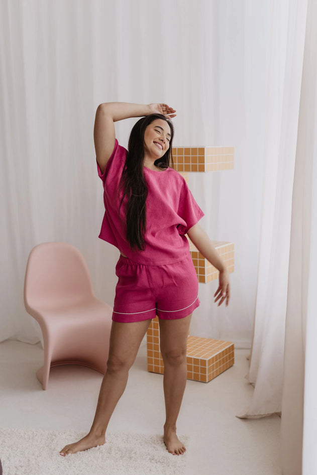 Woman in pink pajama set standing in a room with white curtains and a pink chair.