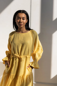Woman wearing a yellow dress standing against a white wall with sunlight casting shadows.