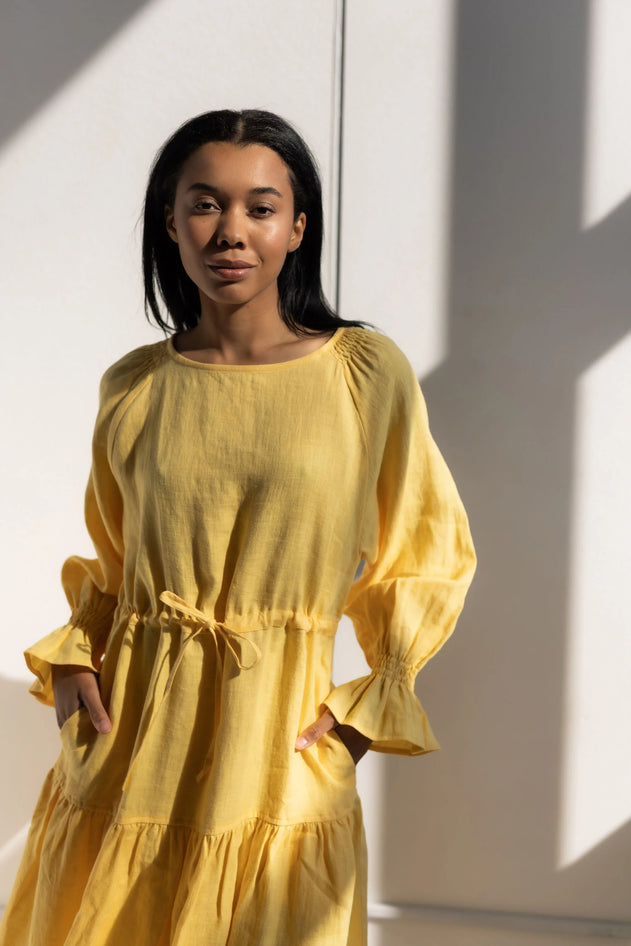 Woman wearing a yellow dress standing against a white wall with sunlight casting shadows.