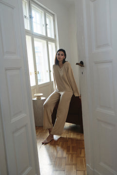 Woman in beige pajamas sitting on a chair by a window in a room with white walls and wooden floor.