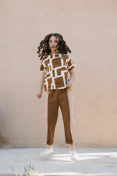 Woman wearing a patterned top and brown pants against a beige wall.