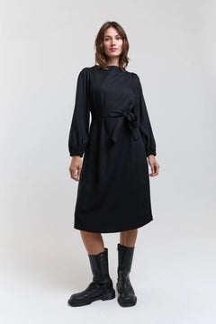 Woman wearing a black dress with long sleeves and a belt, standing against a white background.