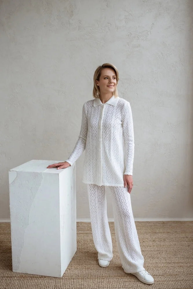 Woman in a white lace outfit standing next to a white cube against a plain wall.