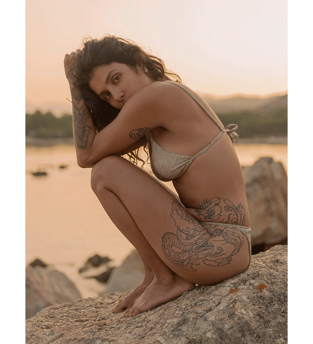 Woman in a bikini sitting on rocks by a body of water at sunset