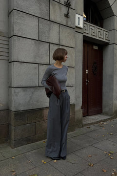 Person standing against a stone building wall holding a brown leather jacket.