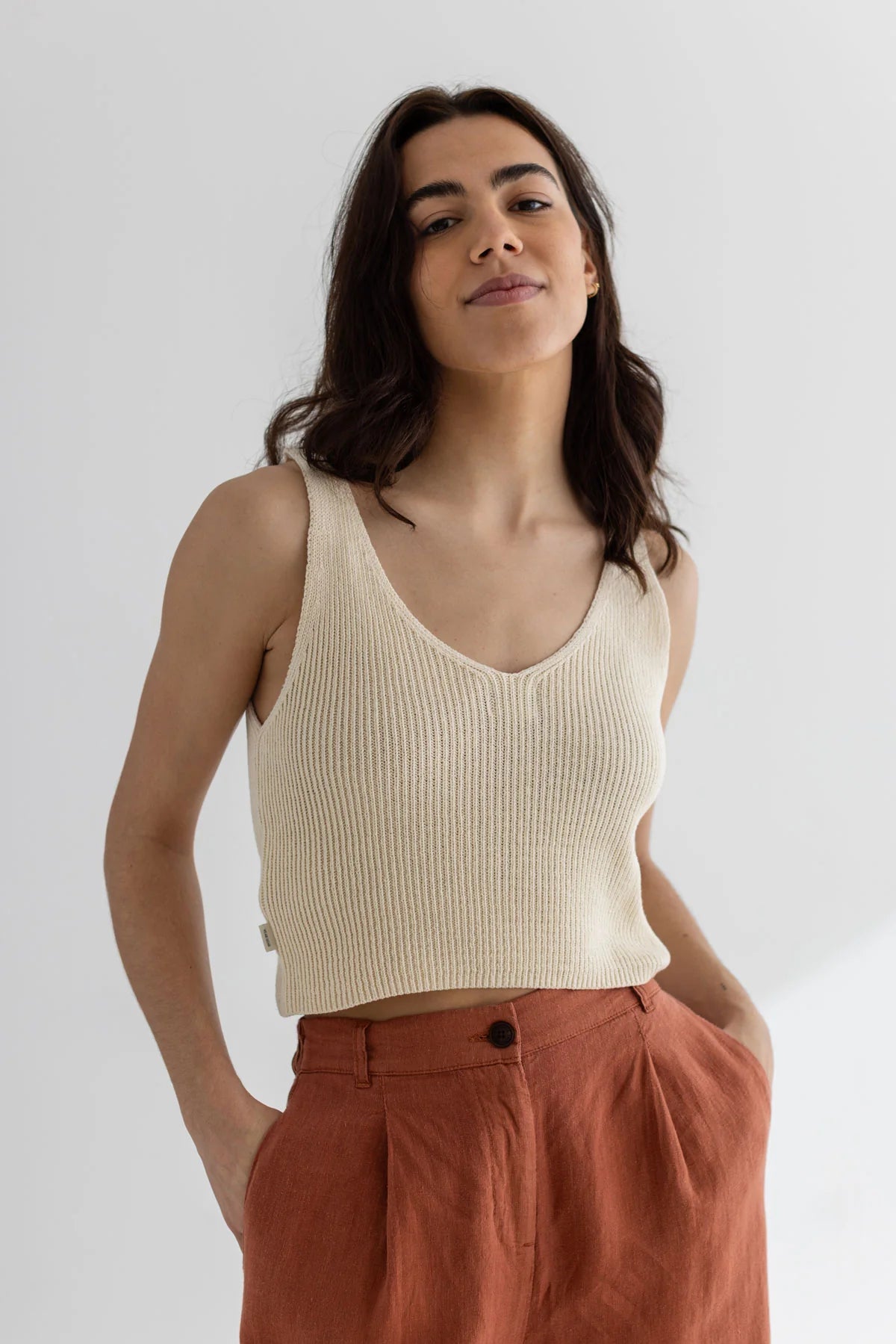 Woman wearing a beige ribbed tank top and rust-colored pants against a white background