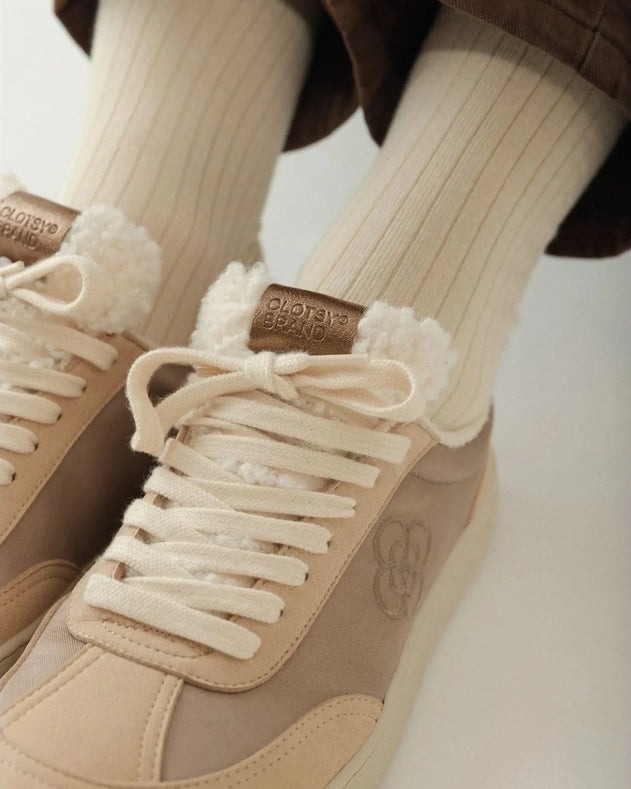 Beige sneakers with white laces and fuzzy accents worn with matching socks on a neutral background