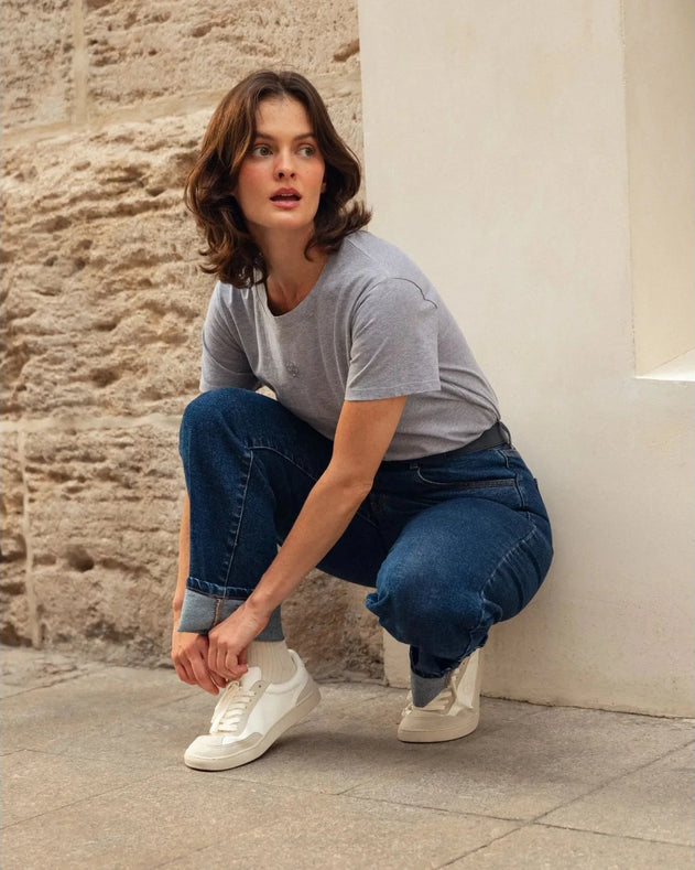 Person wearing a gray t-shirt, blue jeans, and white sneakers against a stone wall.