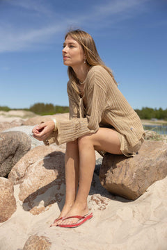 Woman sitting on rocks by a body of water wearing a beige sweater and red sandals.