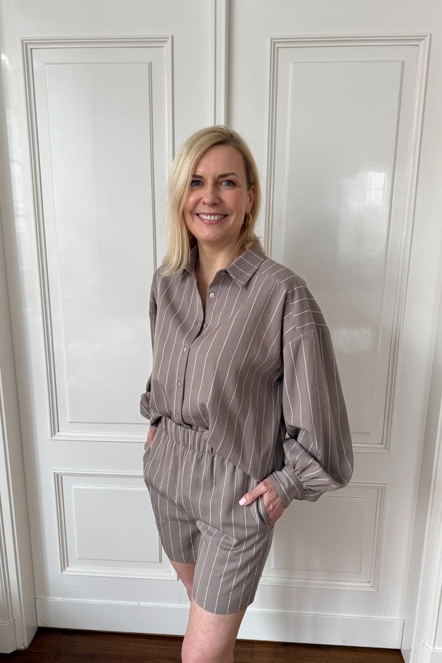 Woman wearing a striped shirt and shorts set against a white paneled wall.