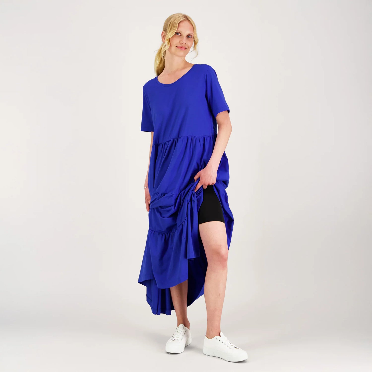 A woman modeling a bright royal blue short-sleeve high-low dress over black shorts and white sneakers, standing against a light gray studio background.