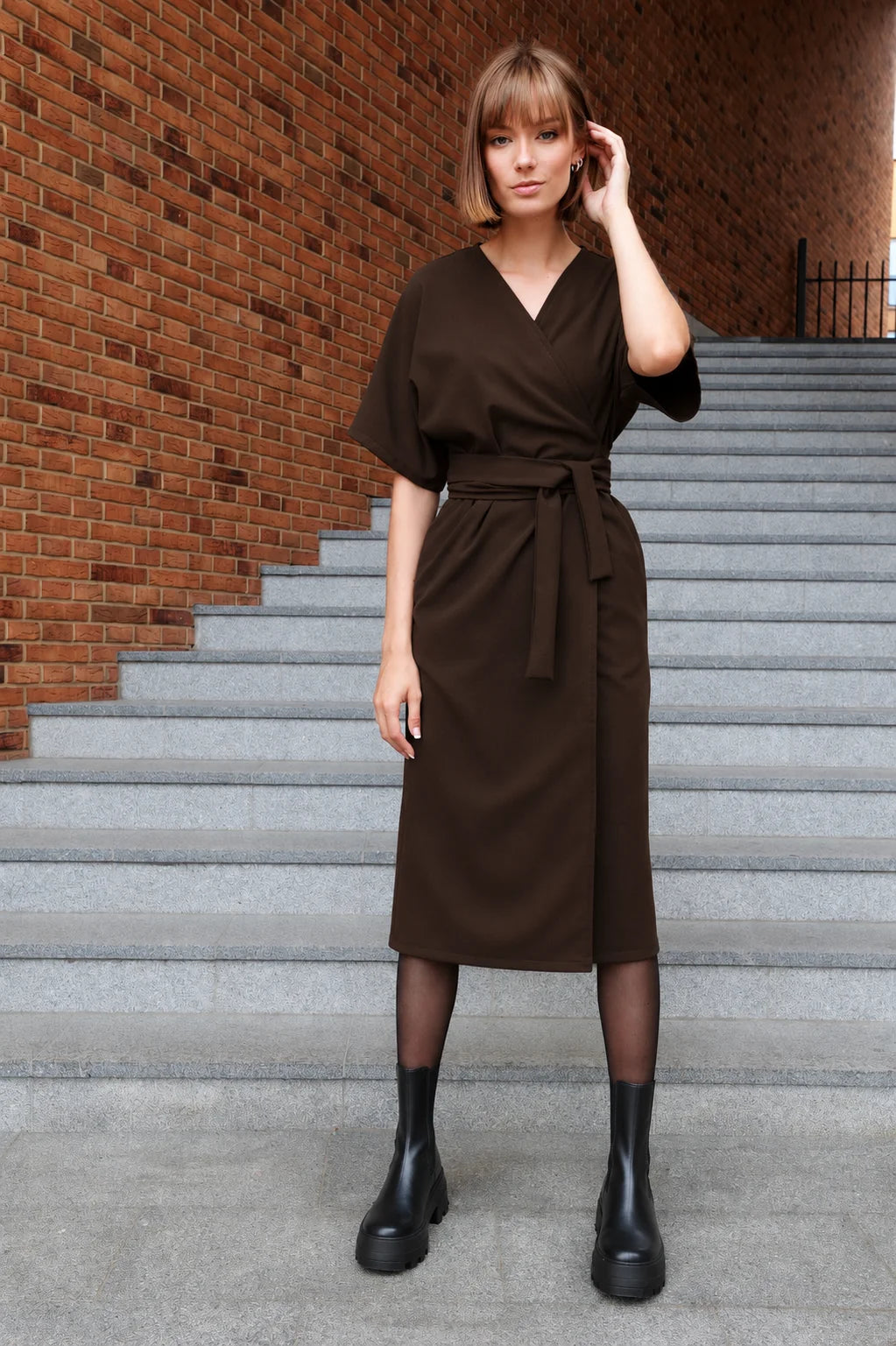 A woman models a dark brown wrap midi dress with short sleeves and a matching tie belt, standing on gray concrete stairs against a red brick wall.