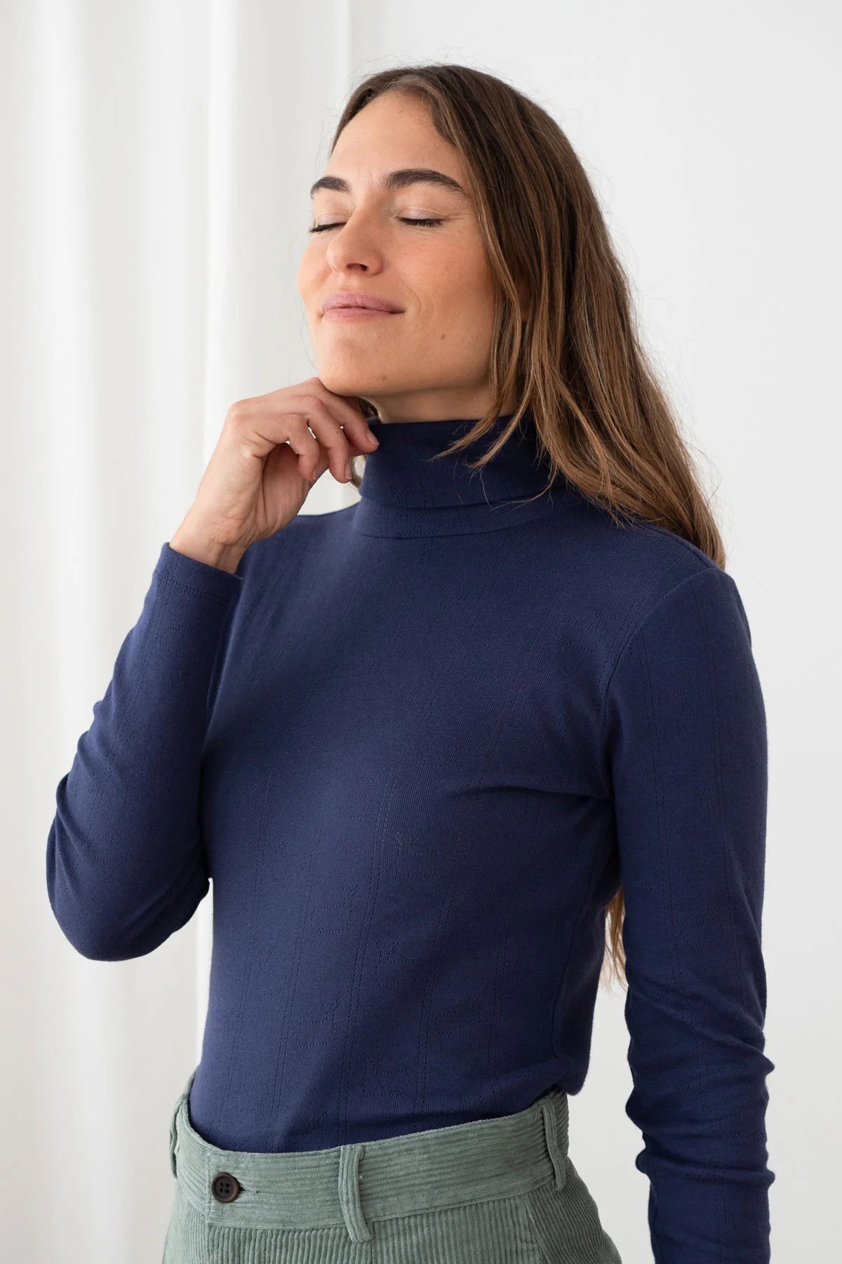 A woman wearing a navy blue ribbed mock neck turtleneck sweater, paired with olive green corduroy pants, posing against a white curtain background.