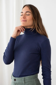 A woman wearing a navy blue ribbed mock neck turtleneck sweater, paired with olive green corduroy pants, posing against a white curtain background.