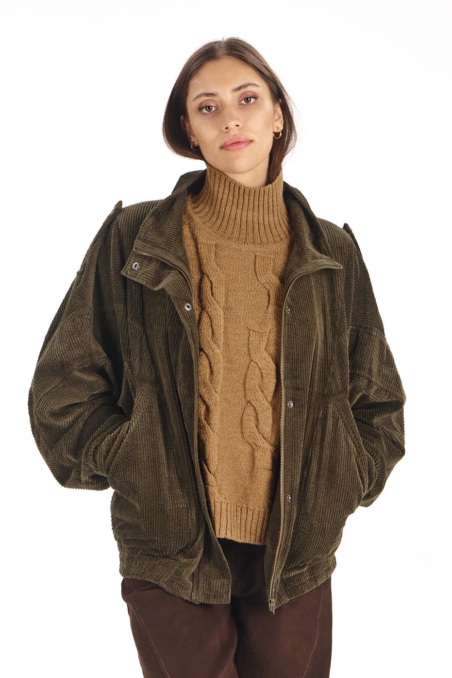 A woman wearing an olive green corduroy jacket over a tan cable-knit turtleneck sweater and dark brown pants, photographed against a plain white background.