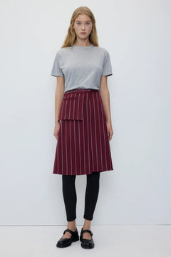 A woman stands against a plain white studio background wearing a light gray short-sleeve t-shirt, a dark red A-line skirt with thin white vertical pinstripes and a front patch pocket, black leggings, and black Mary Jane shoes.