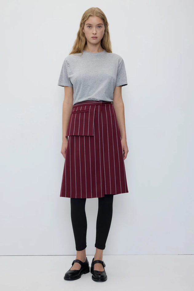 A woman stands against a plain white studio background wearing a light gray short-sleeve t-shirt, a dark red A-line skirt with thin white vertical pinstripes and a front patch pocket, black leggings, and black Mary Jane shoes.