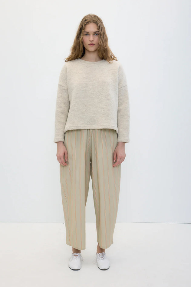 A model stands against a plain white studio background wearing a light beige knit sweater, beige and cream vertically striped wide-leg cropped trousers, and white sneakers.