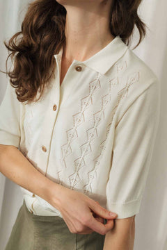 A close-up of a woman wearing a cream-colored short-sleeve knit polo cardigan with a geometric eyelet pattern and brown buttons, paired with olive green trousers.