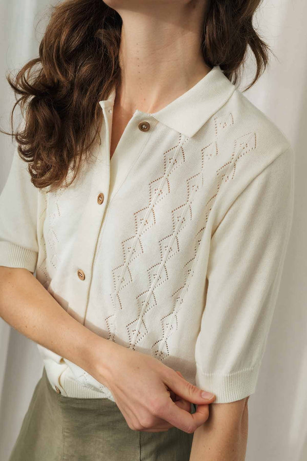 A close-up of a woman wearing a cream-colored short-sleeve knit polo cardigan with a geometric eyelet pattern and brown buttons, paired with olive green trousers.
