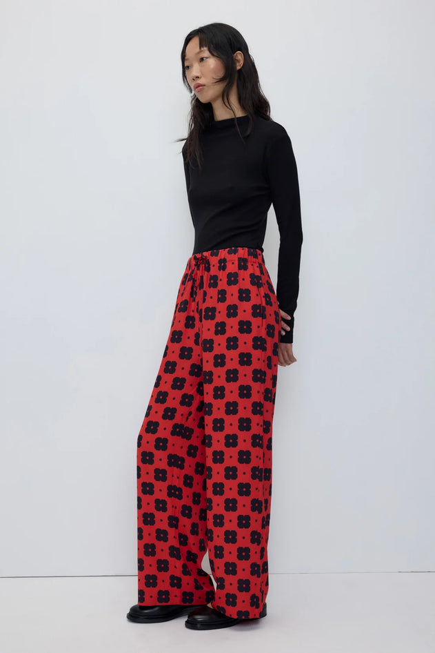 A model stands against a plain white studio background wearing a black long-sleeve mock-neck top and red wide-leg trousers with a black geometric pattern.