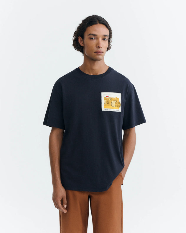 A man wearing a dark navy blue t-shirt with a yellow graphic print on the left chest, paired with brown pants, standing against a light gray background.