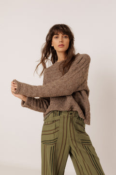 A woman modeling a brown chunky knit sweater and olive green striped trousers against a plain light background.