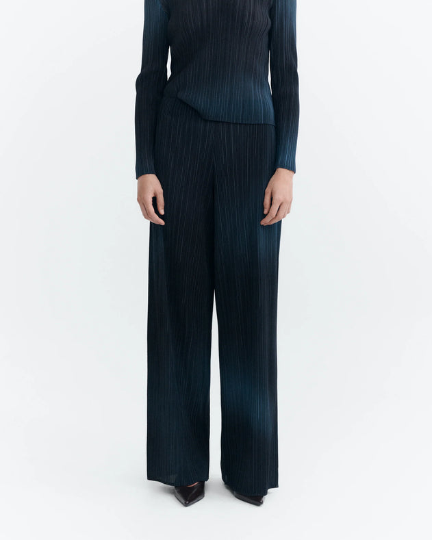 A model wearing a dark blue pleated two-piece set consisting of a long-sleeve top and matching wide-leg trousers, shown from the neck down on a light gray background.