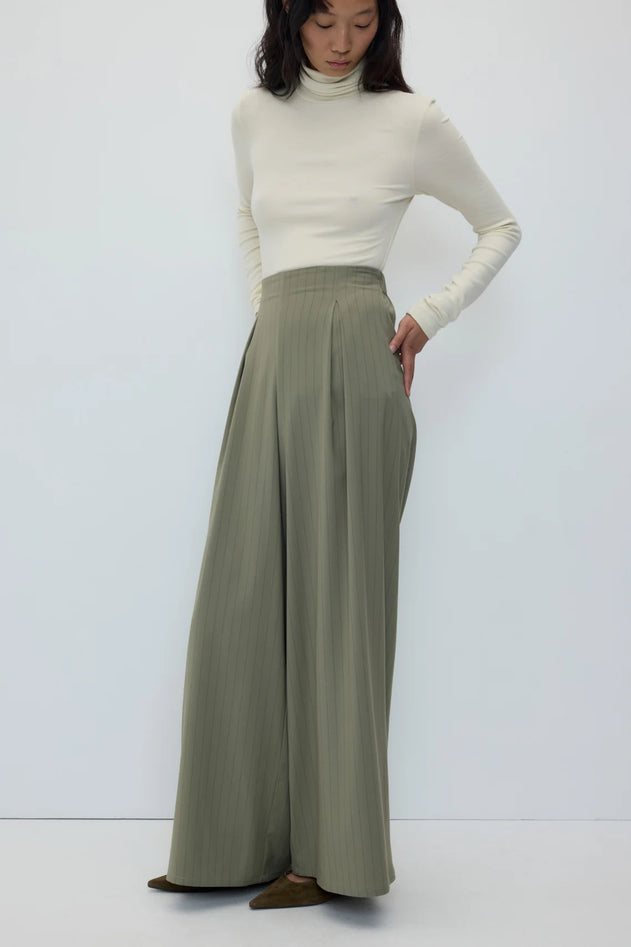 A model wearing a cream ribbed turtleneck sweater and high-waisted, wide-leg sage green pleated trousers, standing against a plain white studio background.