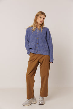 A woman modeling a blue textured button-up cardigan sweater, brown wide-leg trousers, and silver sneakers against a plain light gray studio background.