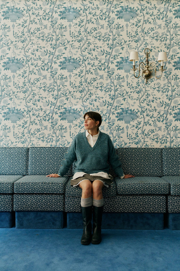 A person sits on a patterned blue sofa against blue floral wallpaper, wearing a teal sweater over a white collared shirt, a short skirt, and tall black boots.