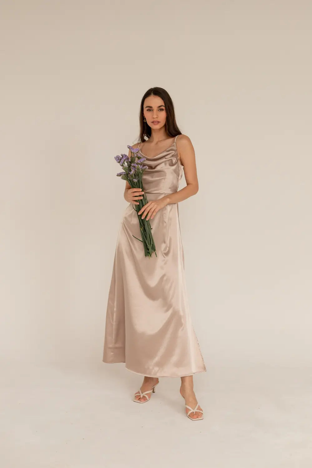 A woman in a beige satin dress with a water neckline and thin straps, holding a bouquet of flowers.